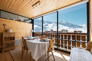 A wooden interior with a table set for dining, a wooden cabinet, and a view of snow-covered mountains through large windows.
