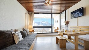 A cozy room with a sofa, wooden table, and benches, a TV mounted on the wall, and a large window offering a mountain view.