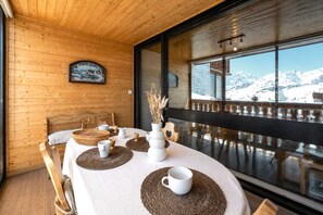 A dining area with a table set for four, wooden walls, a large window with a view of snow-capped mountains, and a framed picture on the wall.