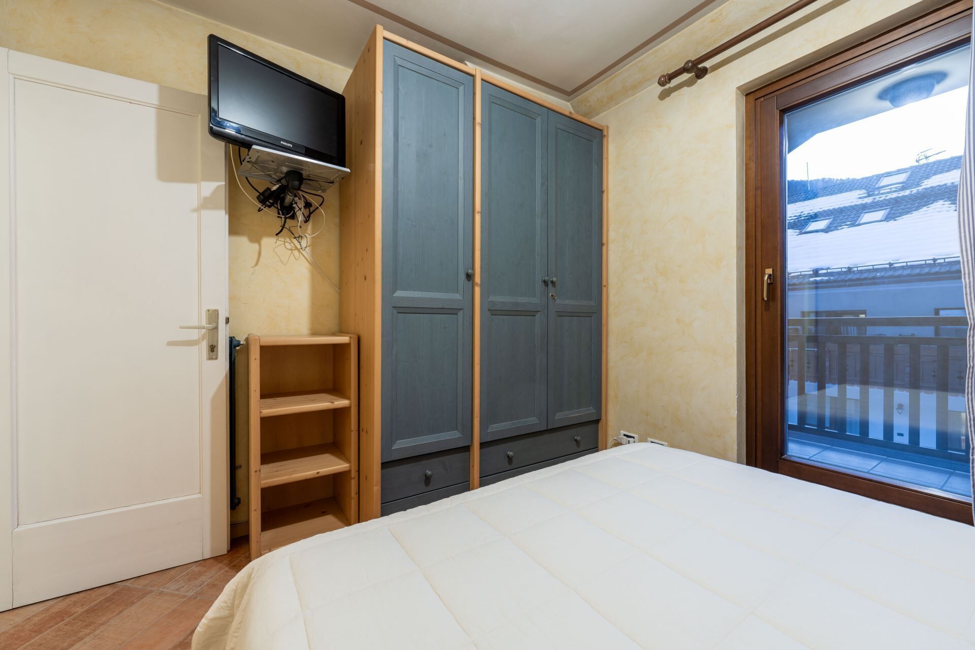 Quarto com cama, guarda-roupa, televisão fixada na parede e varanda com vista.