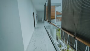 Een gang met witte muren en een betegelde vloer, die uitkomt op een balkon met een metalen hekwerk en uitzicht op gebouwen.