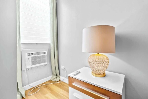 Una habitación con una mesita de noche blanca, una lámpara de pie con base texturizada y una ventana con persianas.