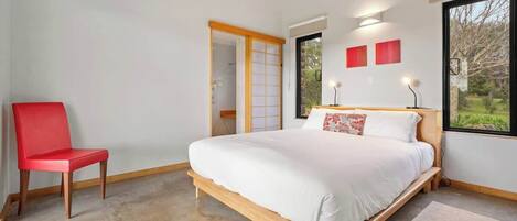 A bedroom with a bed, a red chair, a wooden floor, and a view through a window.