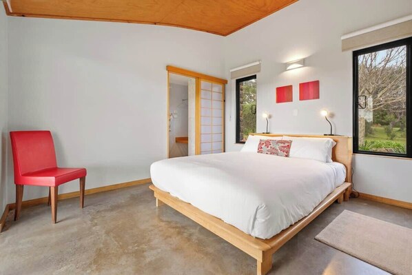 A bedroom with a bed, a red chair, a wooden floor, and a view through a window.