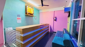A reception area with a reception desk labeled "RECEPTION," a blue bench, a ceiling fan, and a wall-mounted fire extinguisher. - I-ROOMZ S S V Hotel (Hoskote)