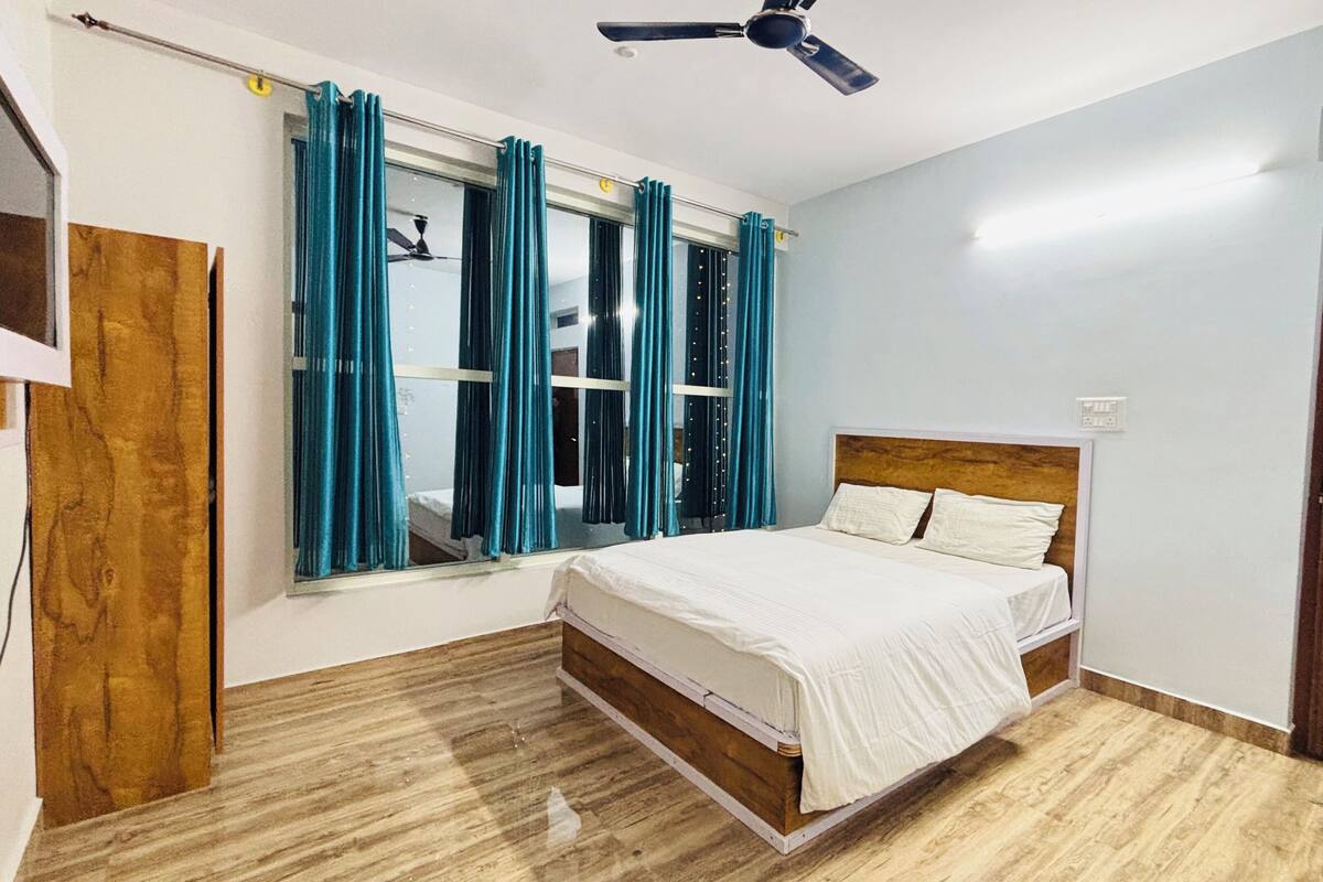A bedroom with a bed, a ceiling fan, a window with curtains, and a wooden door.