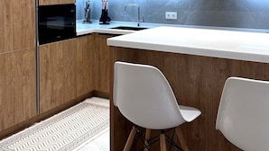 A modern kitchen with wooden cabinets, a white countertop, a built-in oven, and two white bar stools. - Boulevard 46 (Tashkent)
