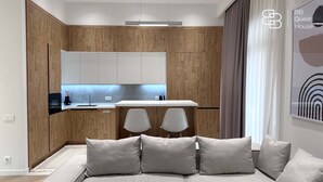 A modern kitchen with wooden cabinets, a white countertop, and a built-in oven. There is a dining area with two white chairs and a table. The room has a neutral color scheme with grey cushions on a sofa. - Boulevard 46 (Tashkent)