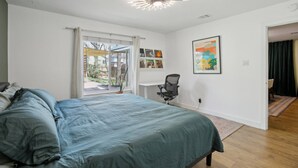 A bedroom with a bed, a desk, and a chair. There is a window with curtains, a ceiling light, and a door leading to another room.