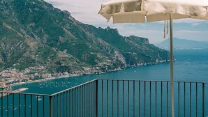 A balcony with a white umbrella overlooking a coastal town with mountains in the background.