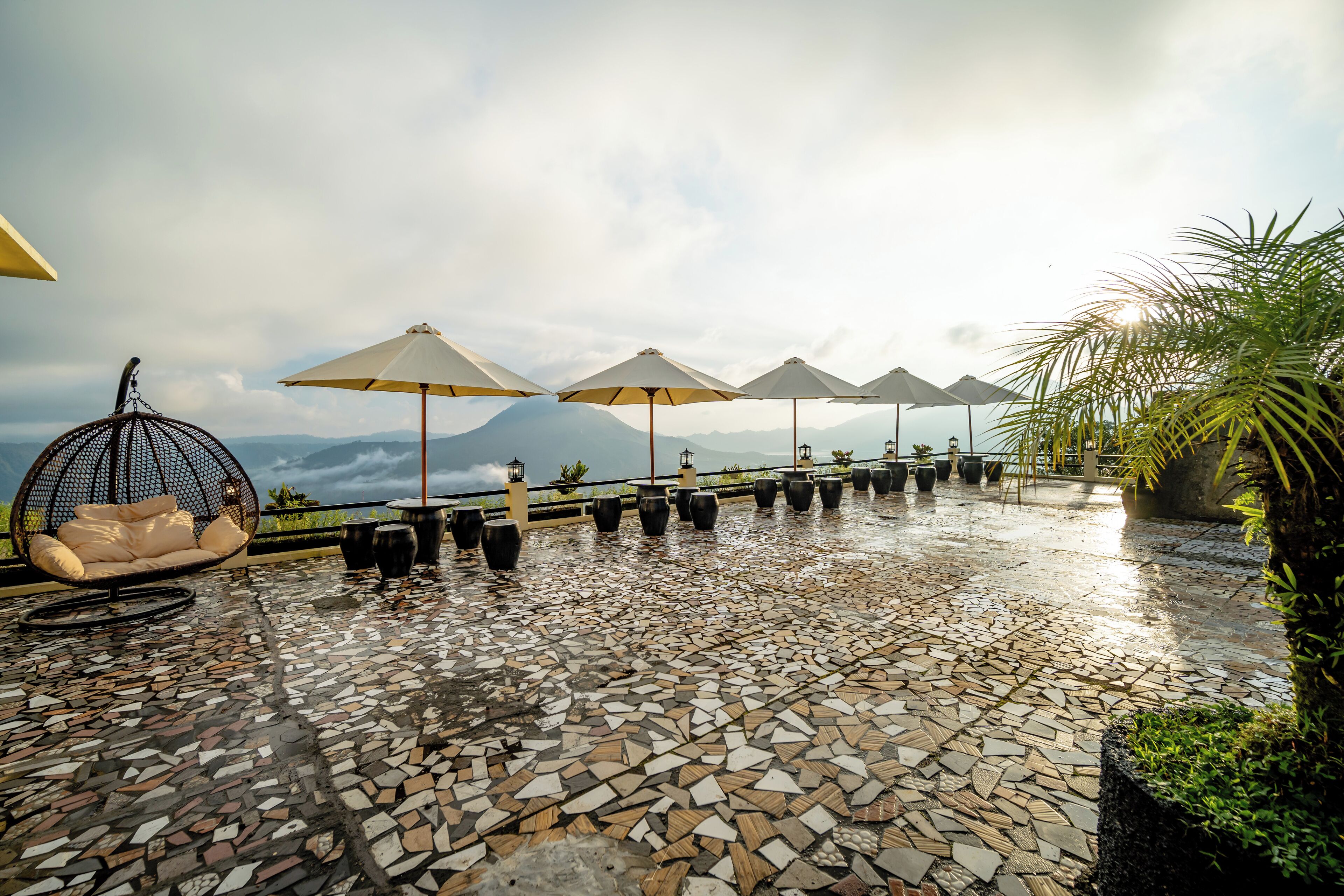 Un patio empedrado mojado con mobiliario exterior, sombrillas y vistas a montañas.