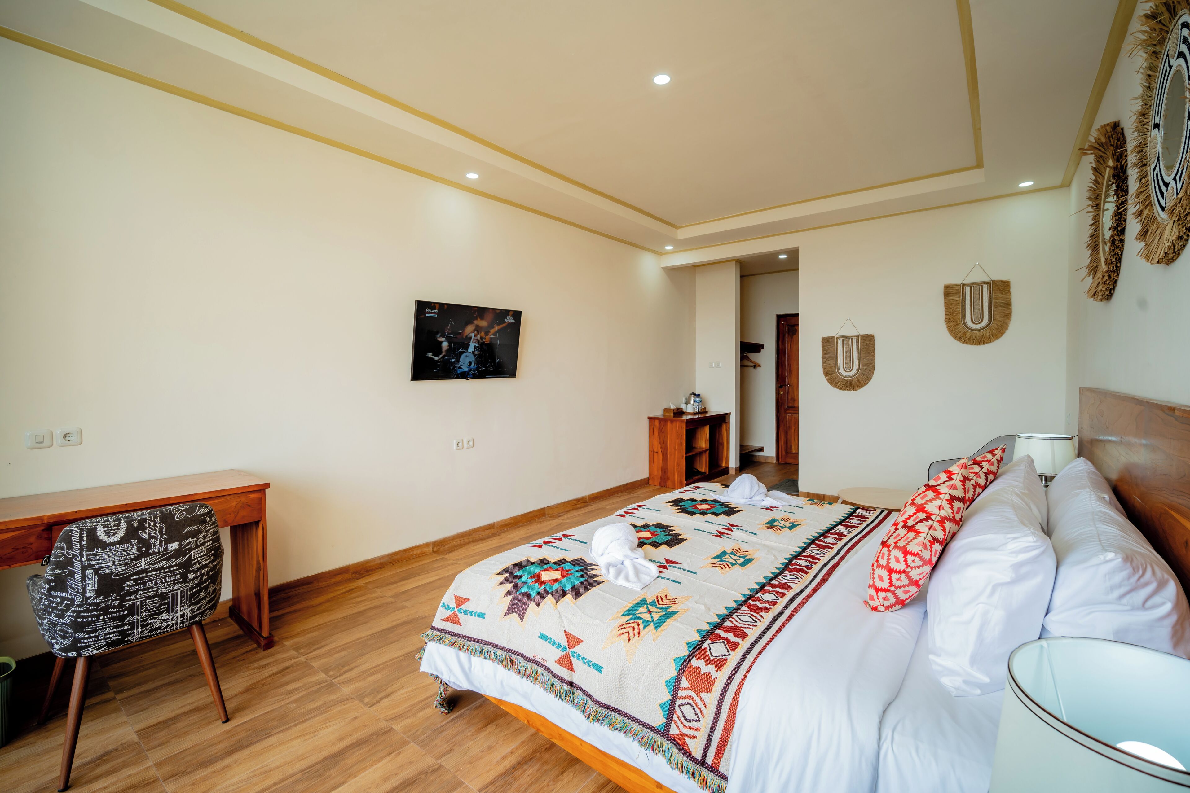 Un dormitorio con escritorio de madera, cama con ropa de cama estampada, televisor montado en la pared y espejo con detalles decorativos.