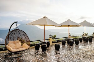 Terraço com cadeira suspensa, mesas ao ar livre e guarda-sóis, com vista para uma paisagem montanhosa.
