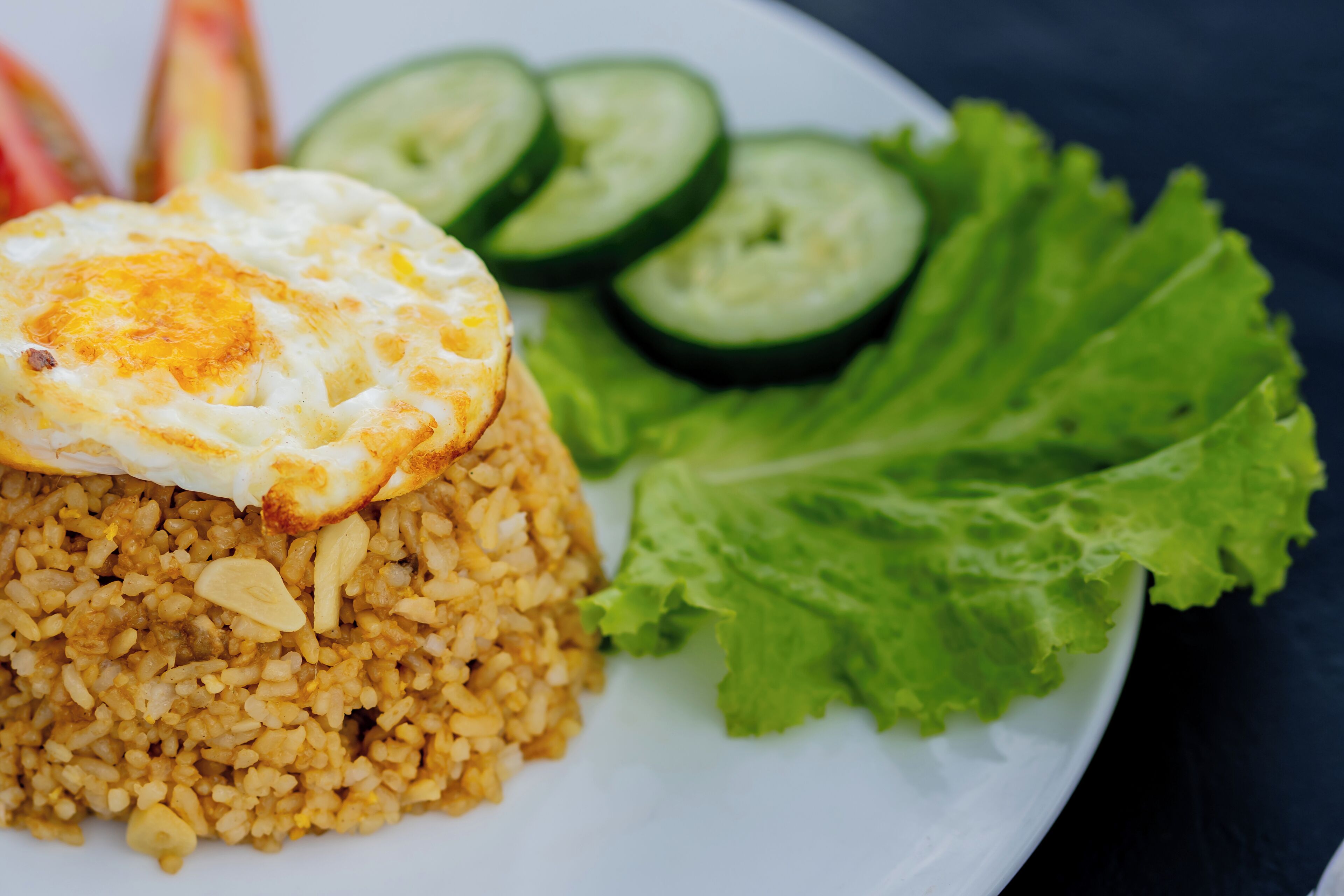 Un plato de arroz frito coronado con un huevo frito, acompañado de rodajas de pepino y lechuga.