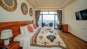 Une chambre d’hôtel avec un grand lit, une tête de lit en bois, une table de chevet avec une lampe, un téléviseur fixé au mur et une vue sur les montagnes par la fenêtre.