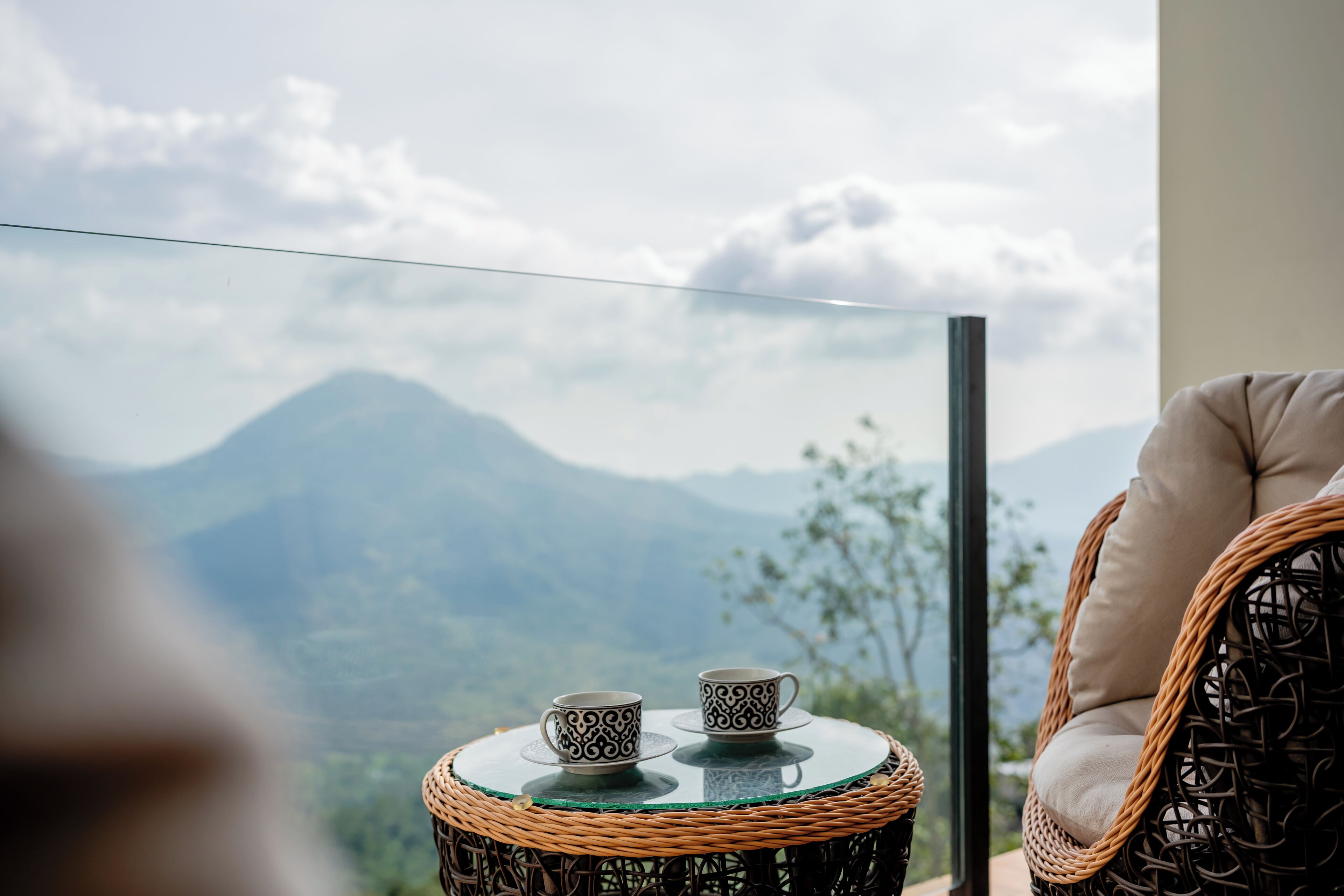 Une chaise en osier et une petite table ronde sur laquelle sont posées deux tasses décorées, le tout offrant une vue sur un paysage montagneux.