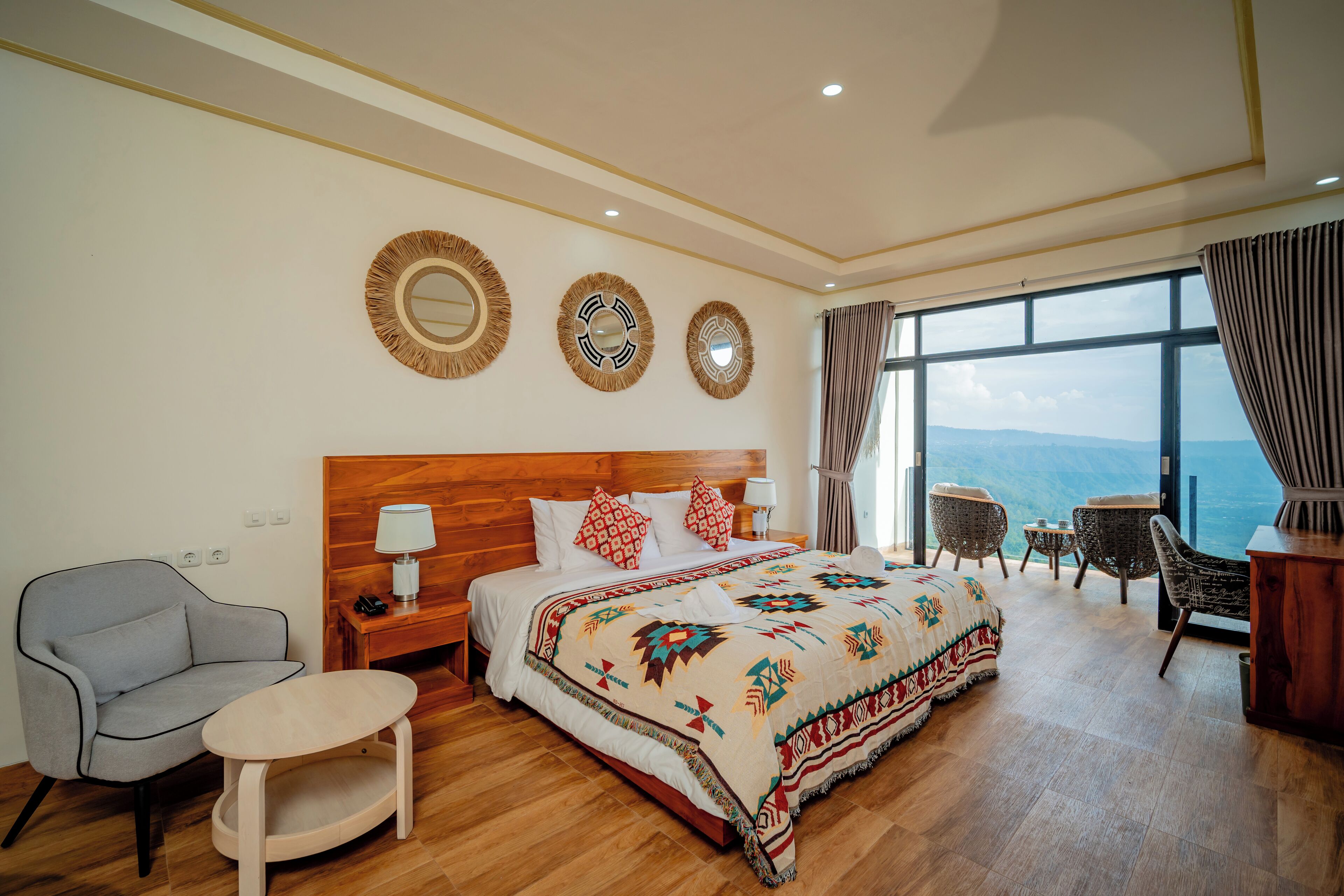 A hotel room with a large bed, a wooden nightstand, a chair, a small table, and a view of the mountains through the window.