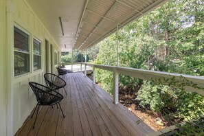 Une véranda couverte avec un plancher en bois, des chaises en métal et une balustrade donnant sur une zone boisée.