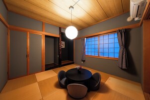 Una habitación tradicional japonesa con suelo de tatami, una mesa redonda con sillas, un ventilador de techo y una ventana con cortinas azules.
