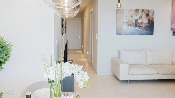 A modern living room with a white sofa, a hanging light fixture, and a painting on the wall. - A bright and cozy Mellieha apartment with modern comforts, close to the beach, shops, and transport—perfect for a relaxing family or couple’s getaway. (Il-Mellieħa)