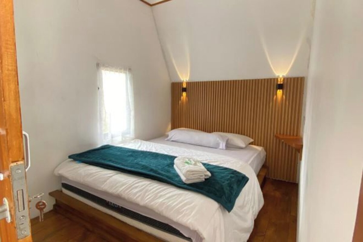 A single bed with a teal blanket and a folded towel, wooden headboard, two wall-mounted lights, and a window with white curtains.
