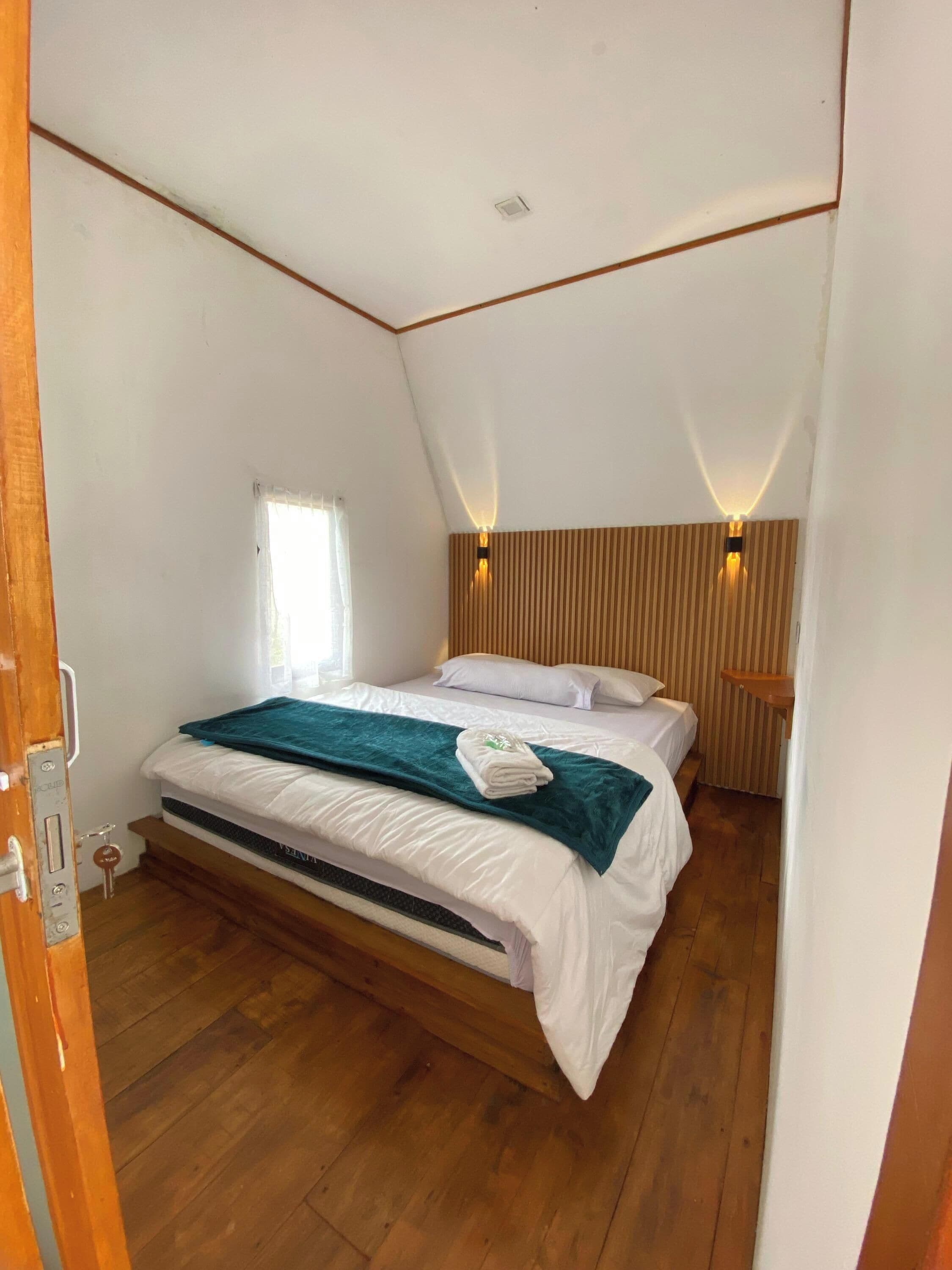 Un dormitorio con una cama individual, suelo de madera, ventana con cortinas y techo inclinado con iluminación empotrada.