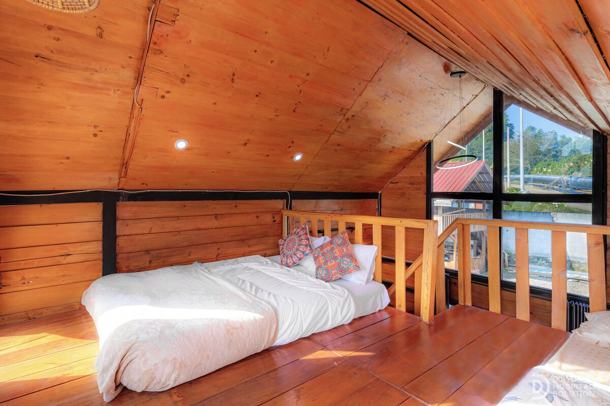 Une pièce de cabane en bois comprenant un lit, une fenêtre et donnant sur un bâtiment aux toits rouges situé à l’extérieur.