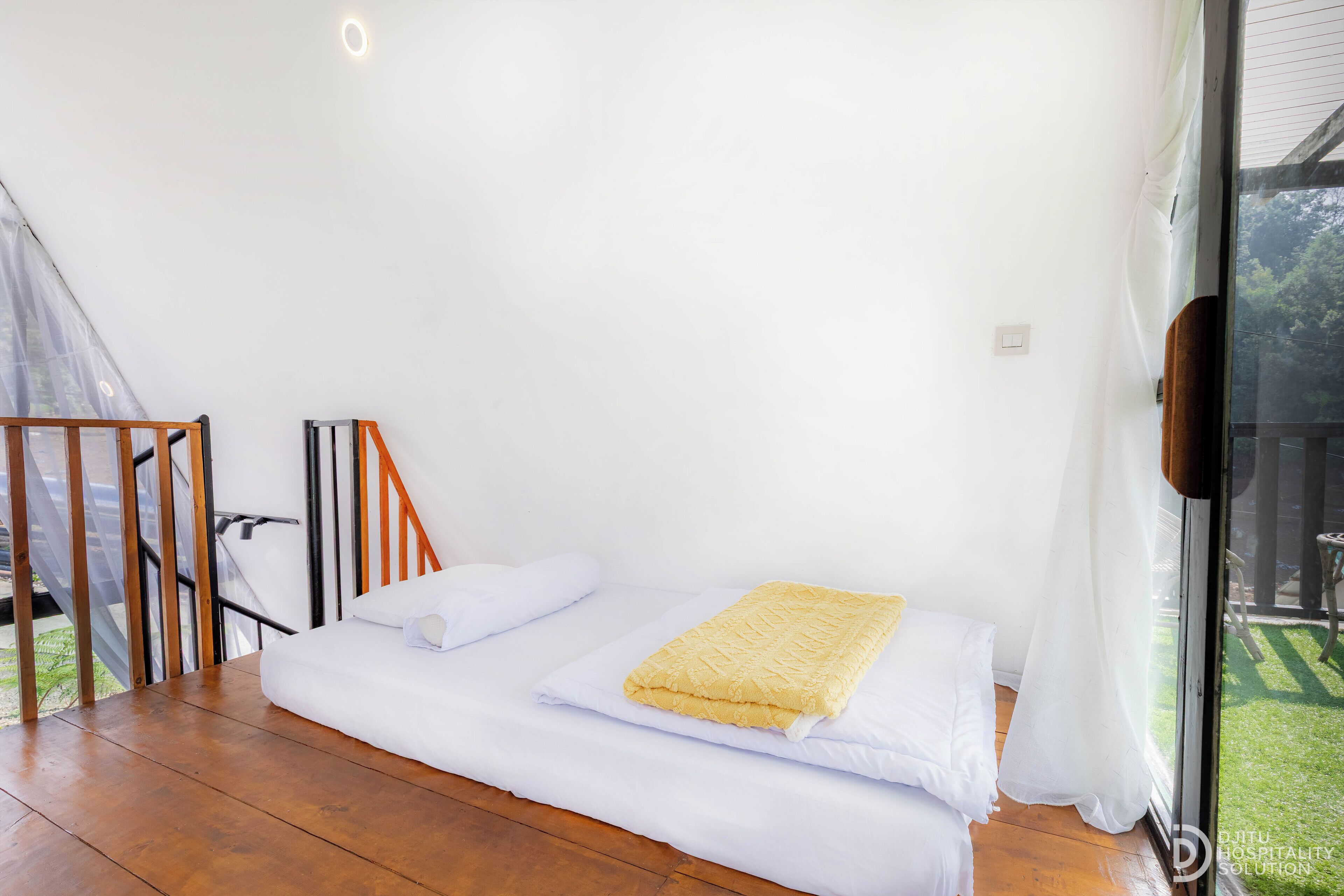 A bedroom with a bed, a wooden floor, a window with curtains, and a view of greenery outside.