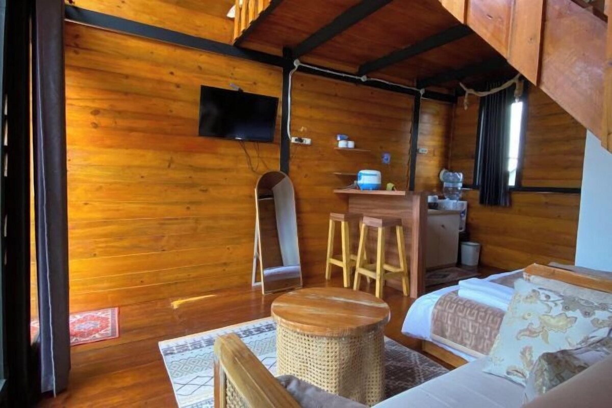 A wooden cabin interior with a bed, a small table, a stool, a television, and a window.