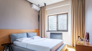 Une chambre d’hôtel avec un lit, une petite table, un radiateur, une fenêtre avec des rideaux et une vue sur les bâtiments environnants.