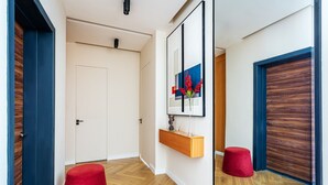 Un couloir avec un plancher en bois, un pouf rouge, une porte en bois, une porte blanche et un miroir orné d’une fleur rouge.