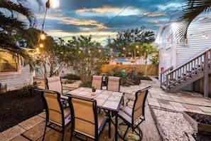 Un patio con mesa y sillas, rodeado de plantas y una terraza de madera.