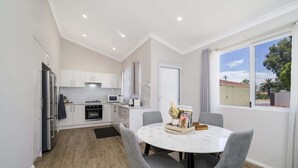 Een moderne keuken met witte kasten, een koelkast en een magnetron. Er staat een eettafel met stoelen en een raam met uitzicht op buiten.