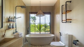 A modern bathroom with a freestanding bathtub, a toilet, and a window with a view of greenery.