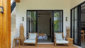 A modern outdoor seating area with two wooden chairs and a small table, leading to a bedroom with a bed and a window.