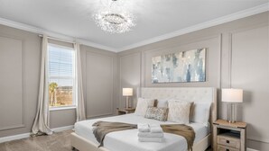 A bedroom with a large bed, two bedside tables, a chandelier, a window with curtains, and a painting on the wall.