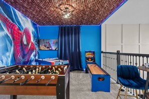 A room with a brick ceiling, a foosball table, a pool table, a Spiderman mural, and a blue wall.