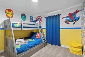 Quarto com beliches, adesivos de super-heróis na parede, uma cortina azul e um pufe amarelo.