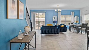 A modern living room with a blue wall, a dark blue sofa, a wooden coffee table, and a dining area with a table and chairs. - Gorgeous 5bed TH close to Disney, Water Park (Davenport)