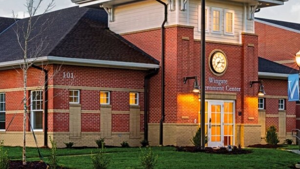 A building with a brick facade, a clock, and a sign reading "Wingate Government Center." - Cozy Bungalow in Wingate w Patio, Grill, Firepit - Easy Access To Bypass & Monroe! +15% Off Stay! (Wingate)