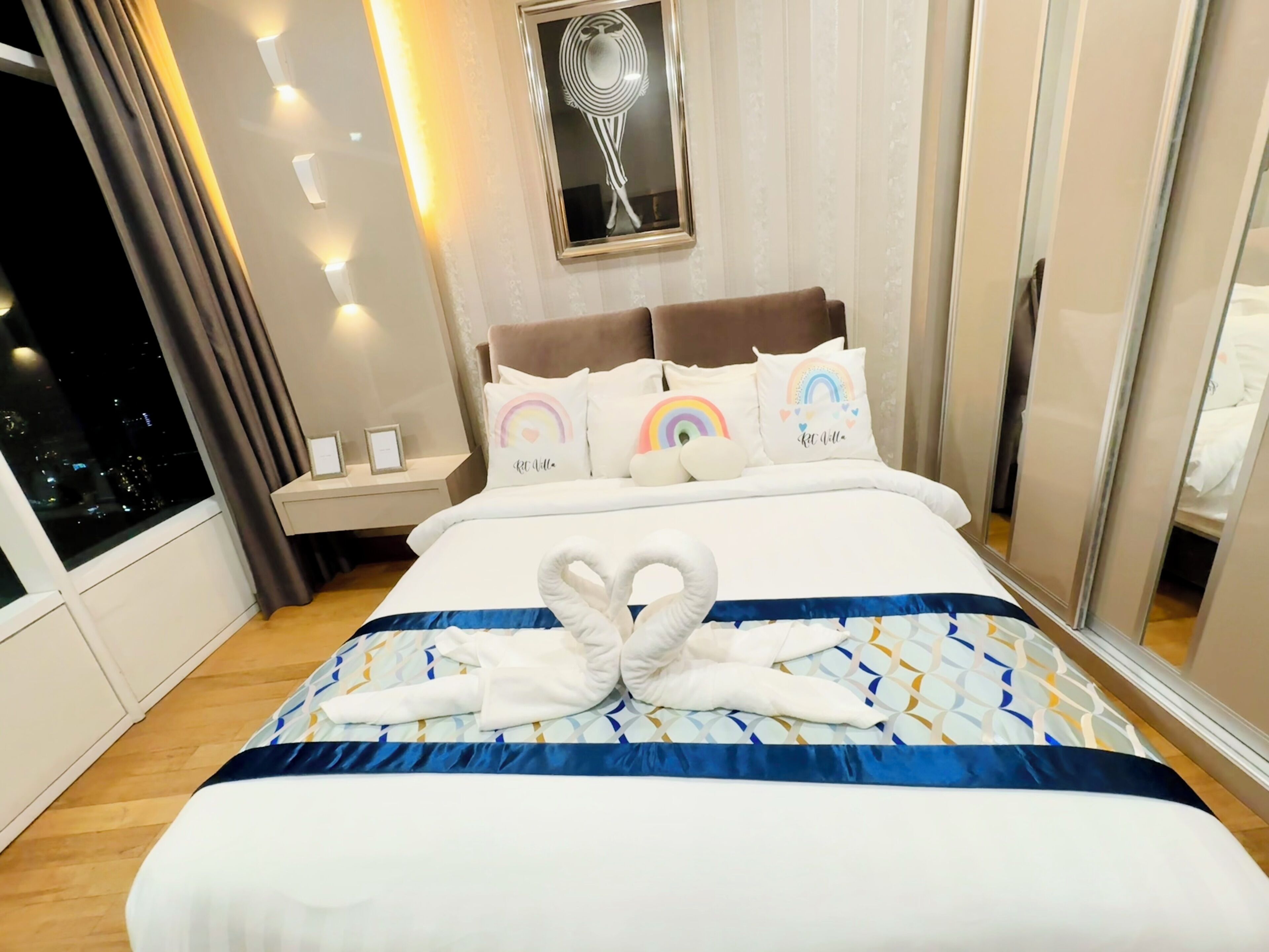 Ein Hotelzimmer mit einem großen Bett, einem Nachttisch, einem gerahmten Kunstwerk über dem Bett und Blick auf die beleuchtete Stadt durch das Fenster.