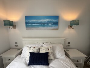 A bedroom with a bed, white nightstands, a white headboard, a beach-themed wall hanging, and two wall-mounted lamps.