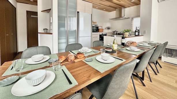 A modern dining area with a wooden table set for six, surrounded by grey chairs. A kitchen is visible in the background with white cabinets and a stainless steel range hood. - ADVENTURe (Furano)