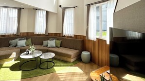 A spacious living room with a large sofa, two coffee tables, and a TV. There are multiple windows allowing natural light and a view of the outdoors. - ADVENTURe (Furano)