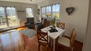 Ein helles Wohnzimmer mit Holzboden, Karoschen Sessel, einem Esstisch mit Stühlen und großen Fenstern mit Vorhängen.
