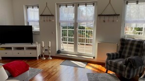 Ein Wohnzimmer mit Flachbildfernseher, einem Karosessel, einer Couch und einem Couchtisch. Zwei Fenster mit Jalousien, eine zur Außenseite führende Tür und eine Heizung sind vorhanden.
