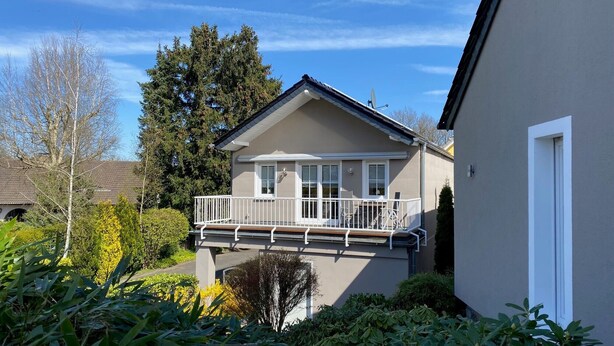 Ein zweistöckiges Haus mit Balkon, umgeben von Grünpflanzen und Bäumen.