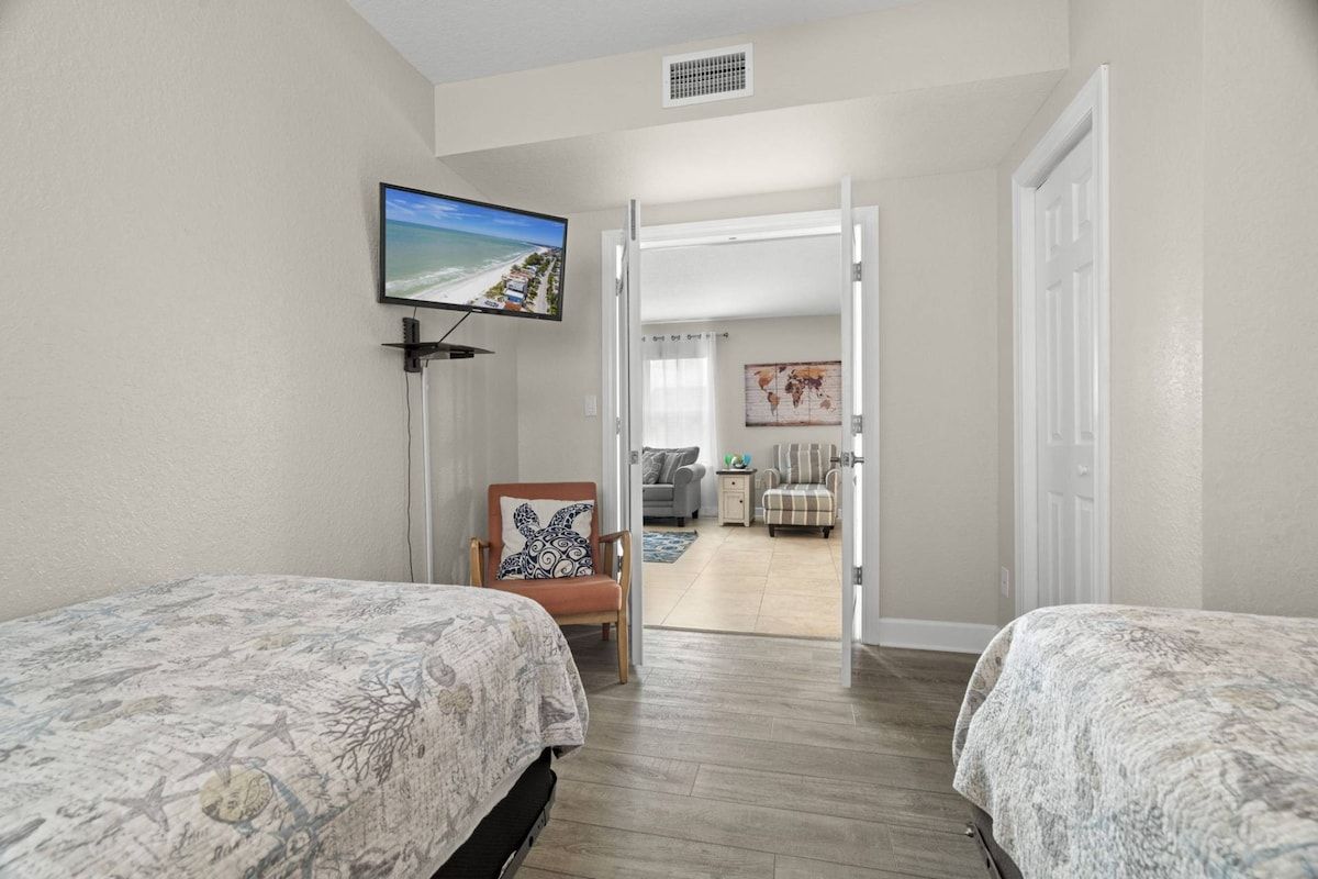 A hotel room with two beds, a television, a chair, and a door leading to another room.