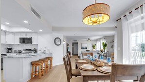 A modern kitchen with white cabinets, a central island with stools, and a dining area with a wooden table set for four. - Updated! Beachside Bi-level Unit, Sleeps 10! FREE Activities! Pool, FREE Parking ~ C400 Sun & Sea (Indian Rocks Beach)