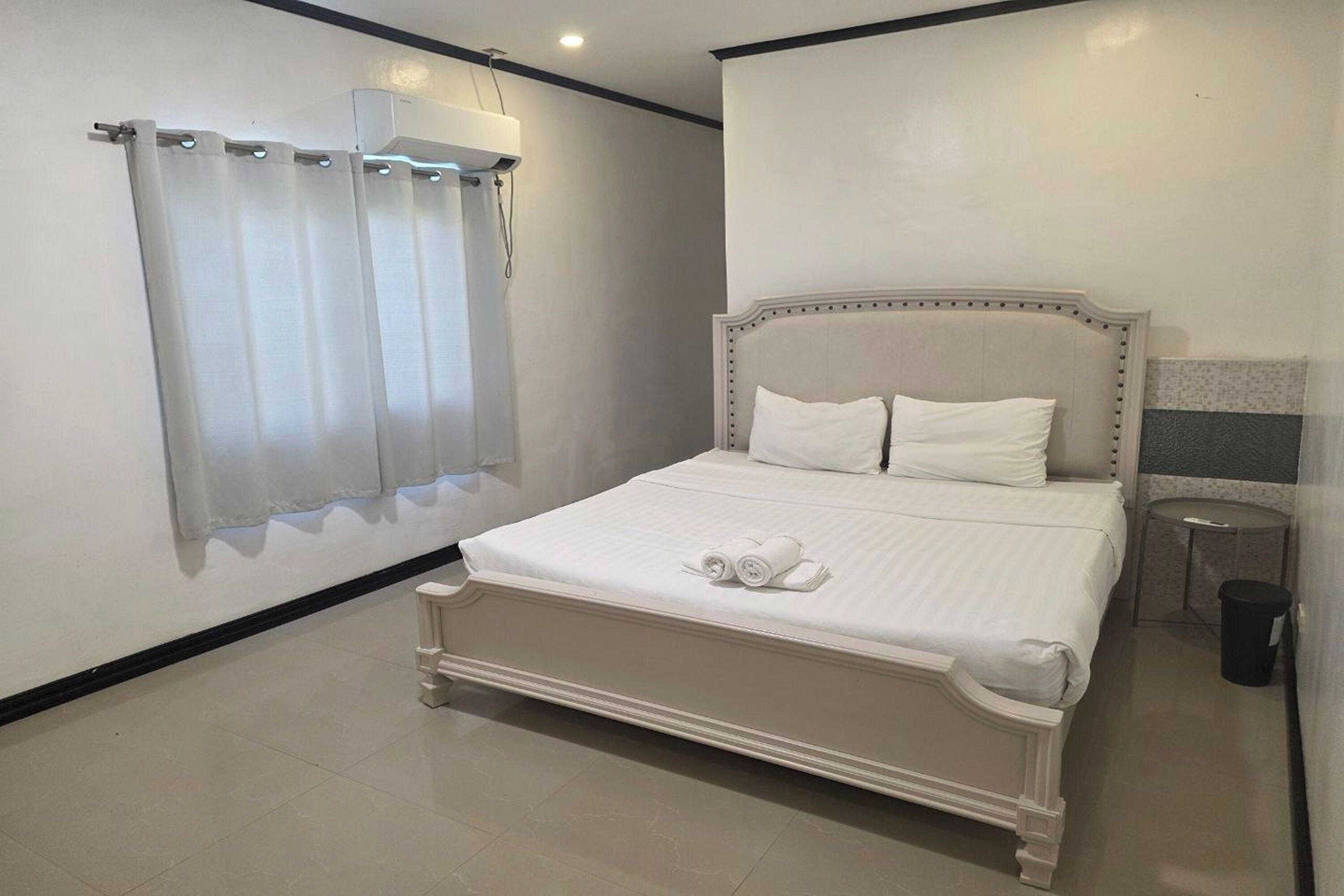 A bedroom with a bed, a window with curtains, a bedside table, and a trash bin.